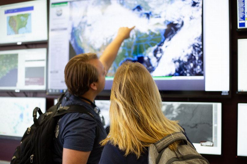 A man and a woman looking at a large tv screen with a weather map of the United States. The man is pointing at a large cloud formation.