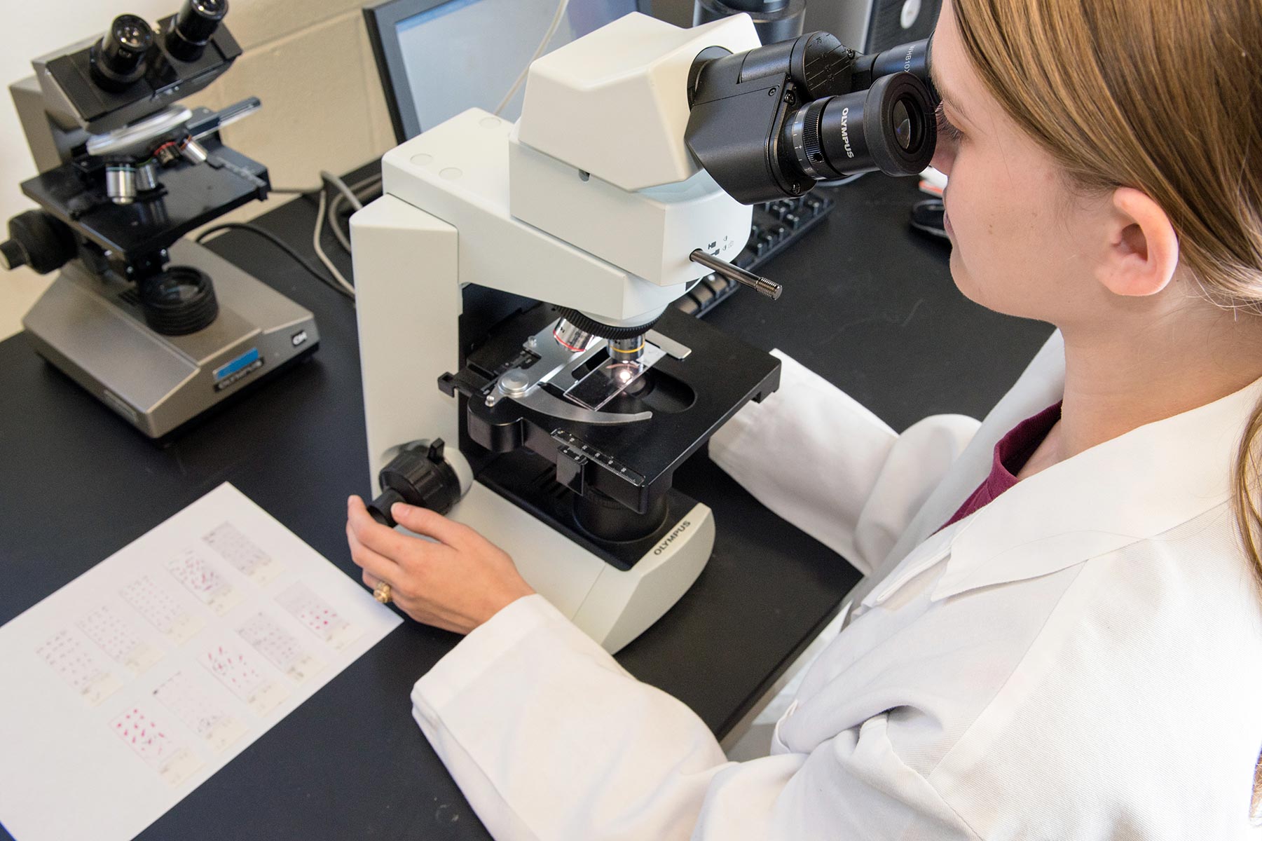 A female student wearing a white lab coat looks into the eyepiece of a microscope within a biology laboratory at Texas A&amp;M University