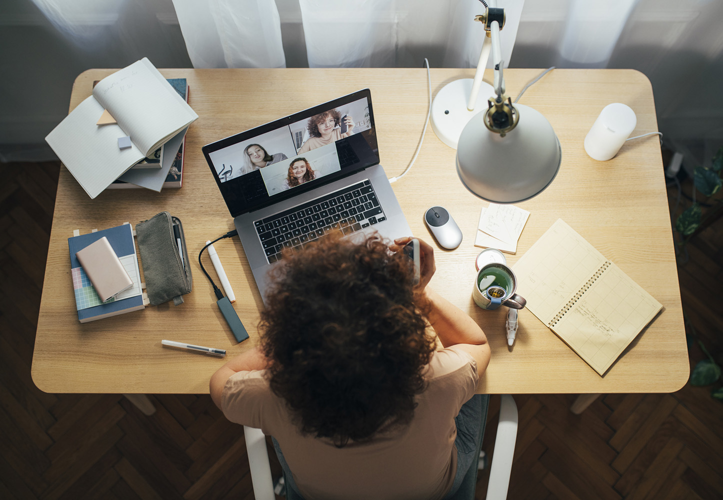 woman sitting at a home desk talking on her cellphone while teleconferencing with colleagues using her laptop computer surrounded by notebooks, a desk lamp and a coffee cup