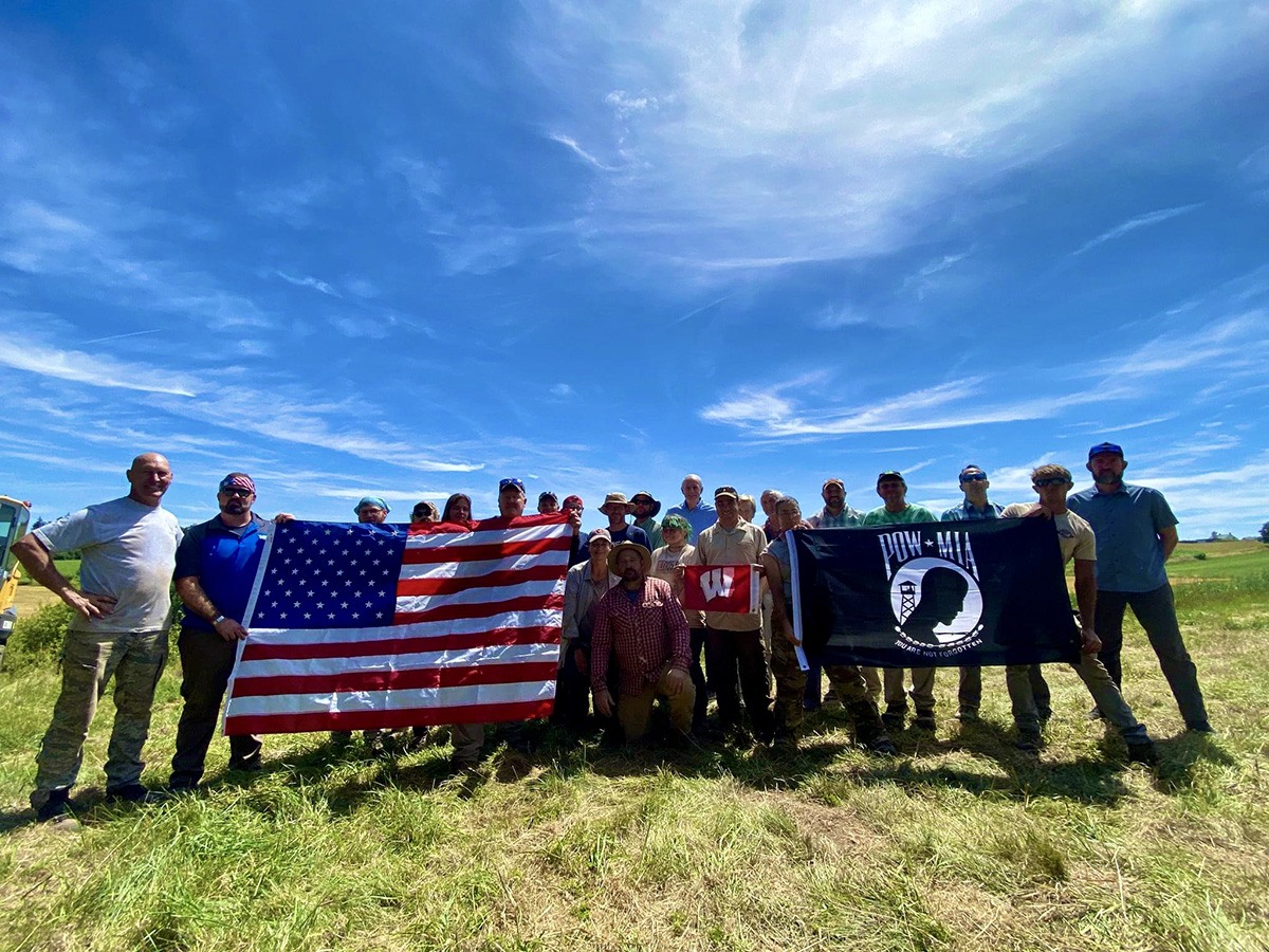 A group of student and faculty pose with an American flag and a POW-MIA flag during a dig in Belgium