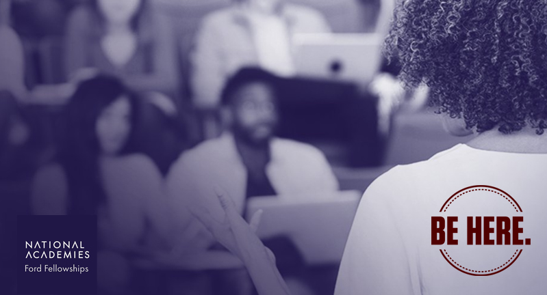 A female presenter with outstretched left hand addresses a multi-race and gendered audience seated in lecture-style classroom