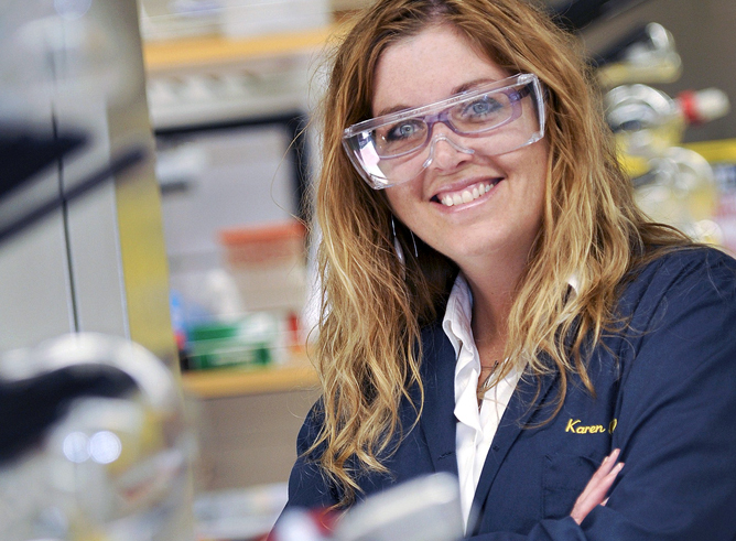 Texas A&amp;M chemist Karen Wooley smiles for the camera in her laboratory while wearing goggles and her trademark blue labcoat