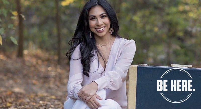 Texas A&M women's and gender studies major Odyssey Olmos smiles for the camera while sitting beside two suitcases in a tree-lined background setting