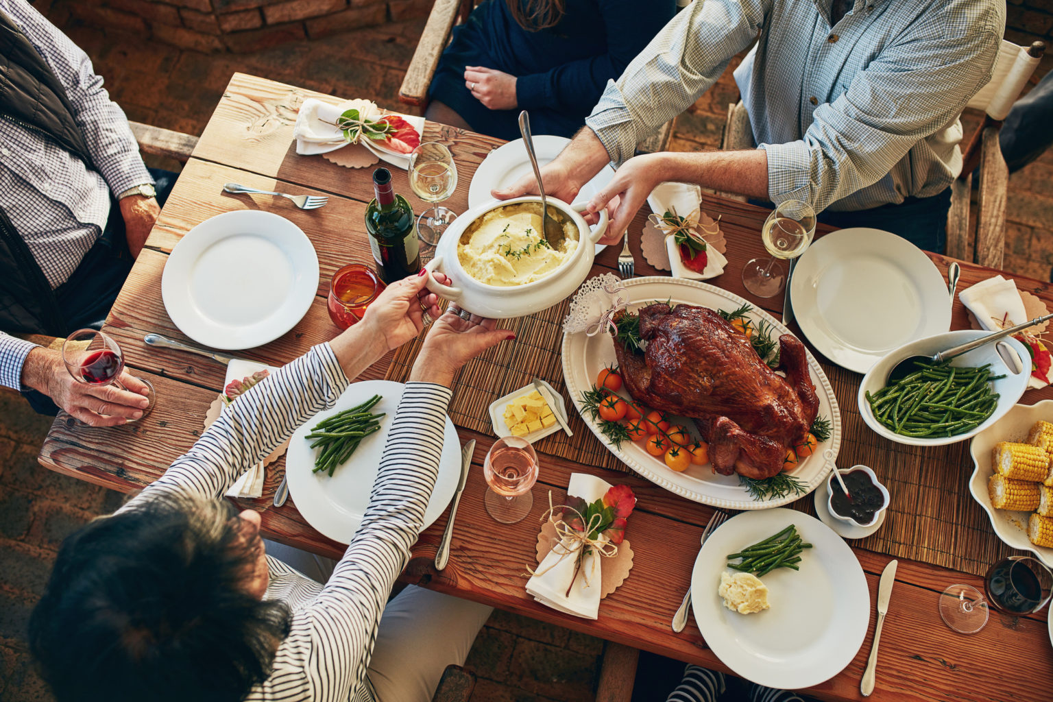 Overhead view of a man and a woman passing the potatoes while sitting down at a wooden table adorned with elegant place settings for a family style meal featuring turkey, potatoes and gravy, green beans and other sides