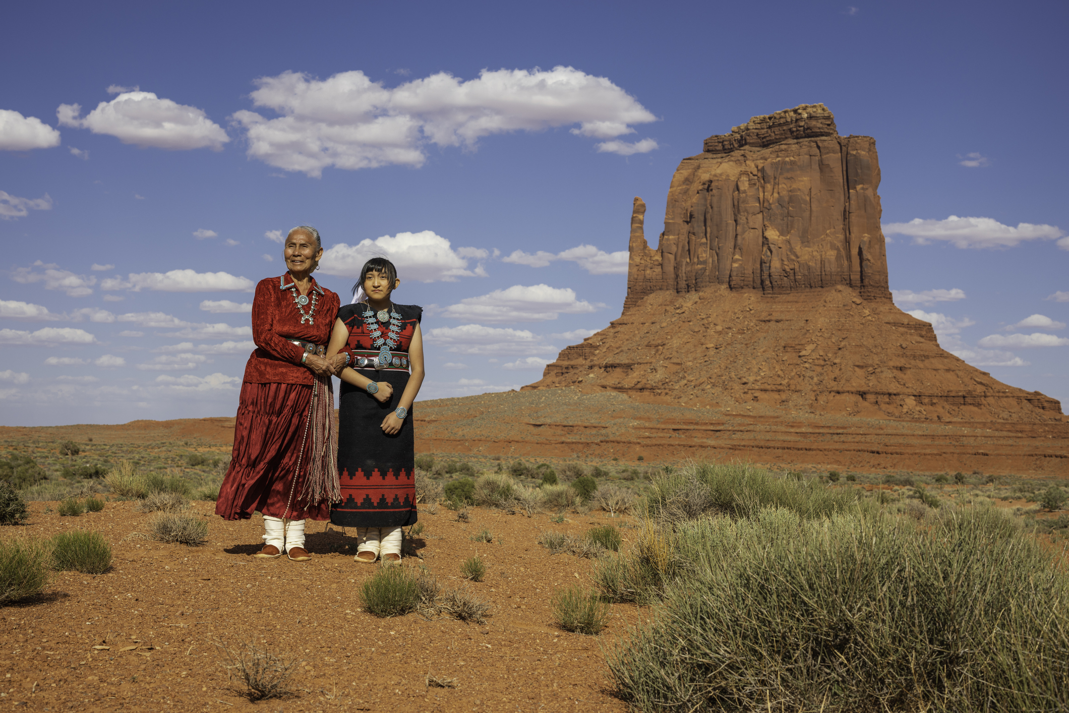 Two individuals in traditional dress stand in front of Merrick Butte in Monument Valley