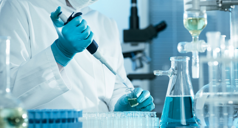 Stock image of a researcher wearing blue latex gloves while pipetting at a laboratory bench filled with glassware, microscopes and other research-related equipment