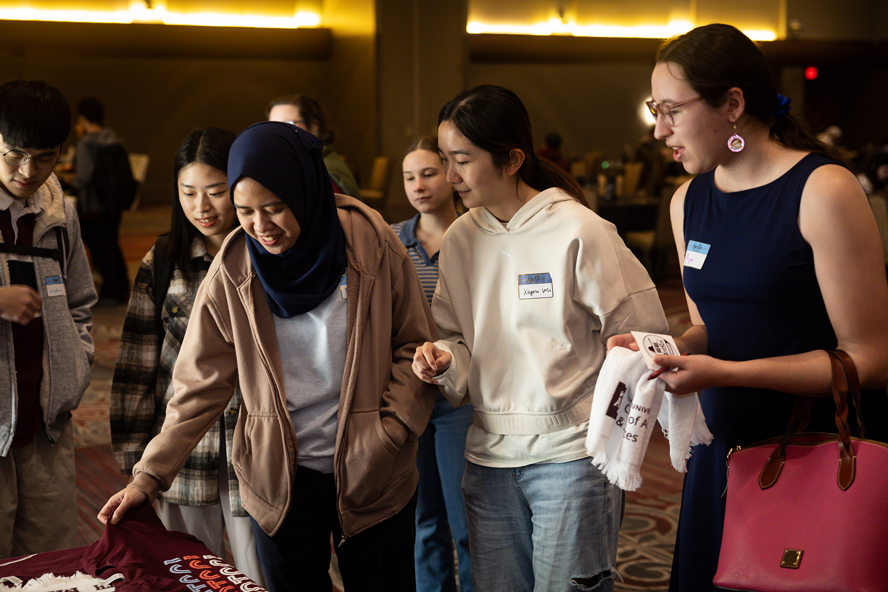 Group of attendees gather around the merchendise table, examining materials on a table. One individual is wearing a hijab.