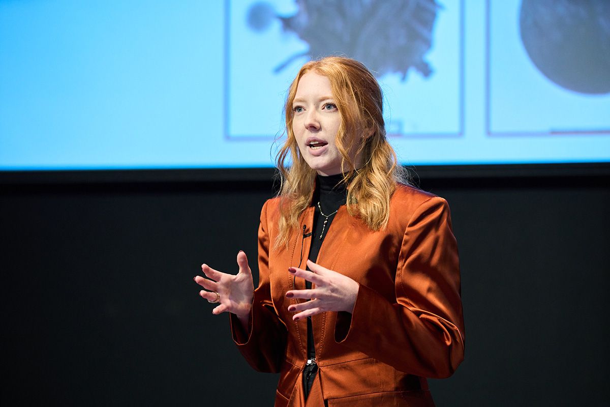 A speaker in an orange blazer is presenting at a TEDx event, gesturing with hands, with a projected image in the background.