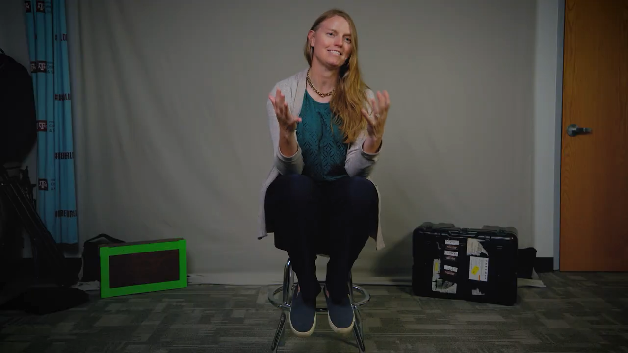 Dr. Jessi Halligan itting on a stool, clapping hands with a joyful expression, in a room with photography equipment visible in the background.