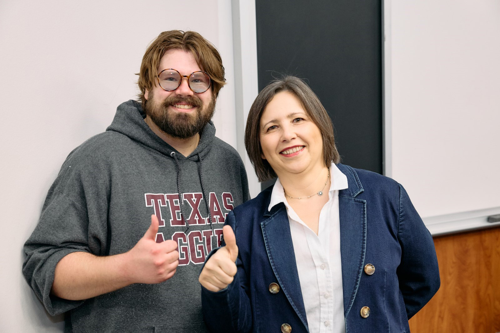 Jeremy in a Texas A&amp;M sweatshirt and Oksana in a navy blazer, standing in a classroom with their thumbs up.