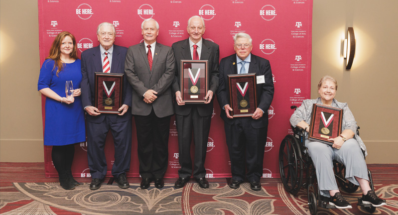 Group of six individuals holding awards, standing in front of a Texas A&M University banner with the slogan "Be Here.