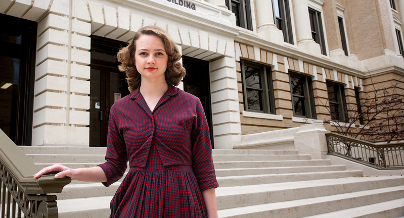 An individual standing confidently on the steps in front of the Melbern G. Glasscock Building, wearing a burgundy dress and a serious expression.