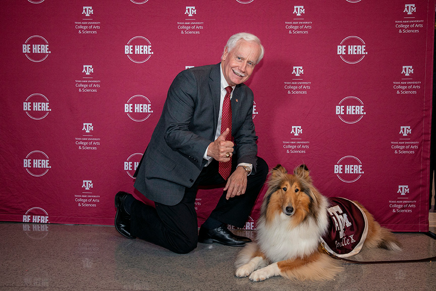Person kneeling next to Reveille, the Texas A&amp;M University mascot, in front of a backdrop with the university's logos and the phrase "Be Here." Both are looking at the camera.