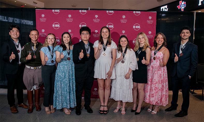 Group of ten Texas A&amp;M University students displaying thumbs-up at an event, standing in front of banners with university logos.